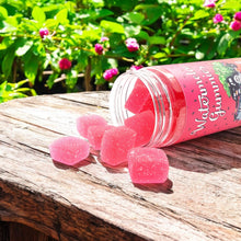 Load image into Gallery viewer, A clear jar with pink watermelon CBD gummies spilling out onto a wooden surface, with greenery in the background.